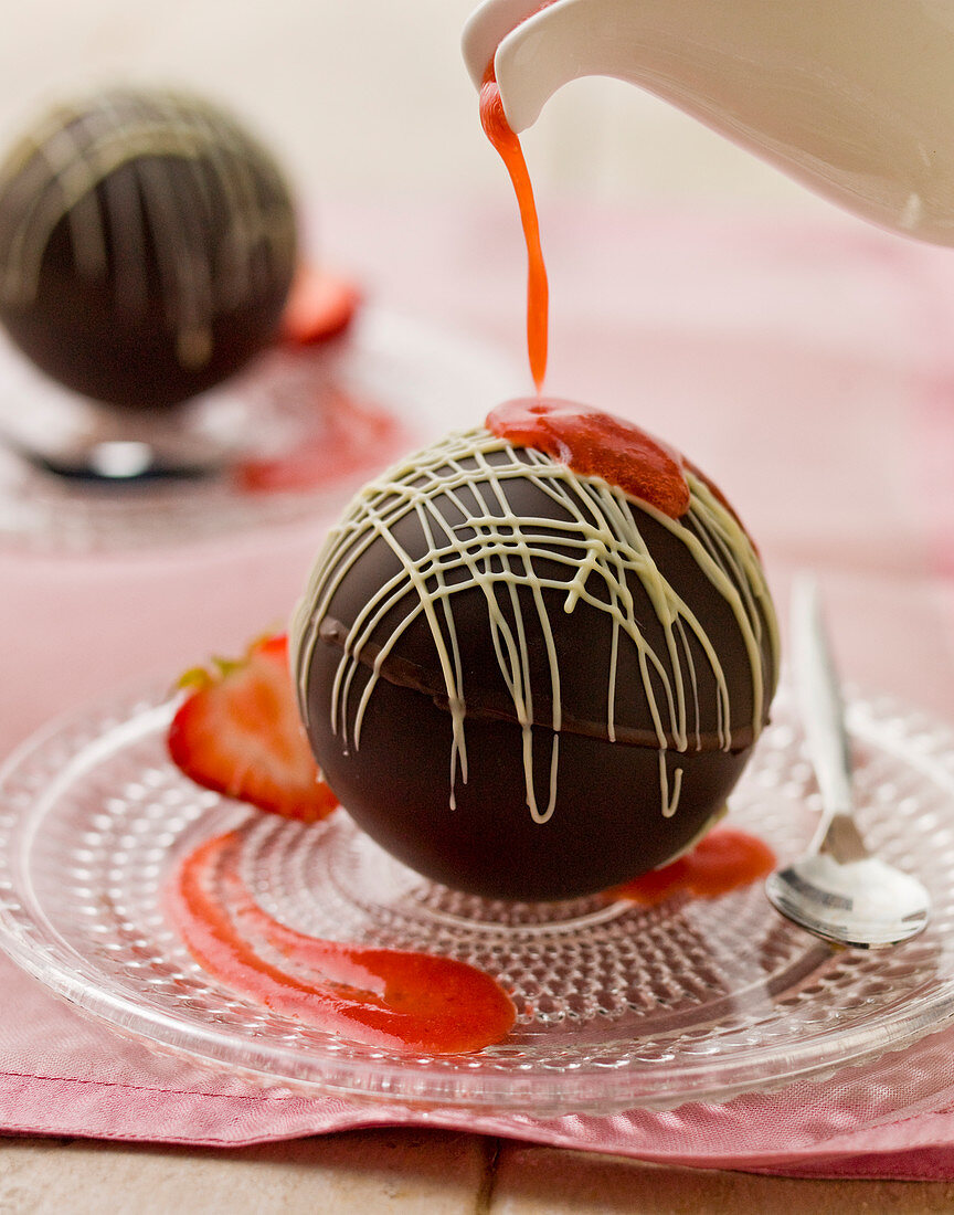 Chocolate spheres with melting hot strawberry sauce being poured from a white jug, on a glass plate and spoon