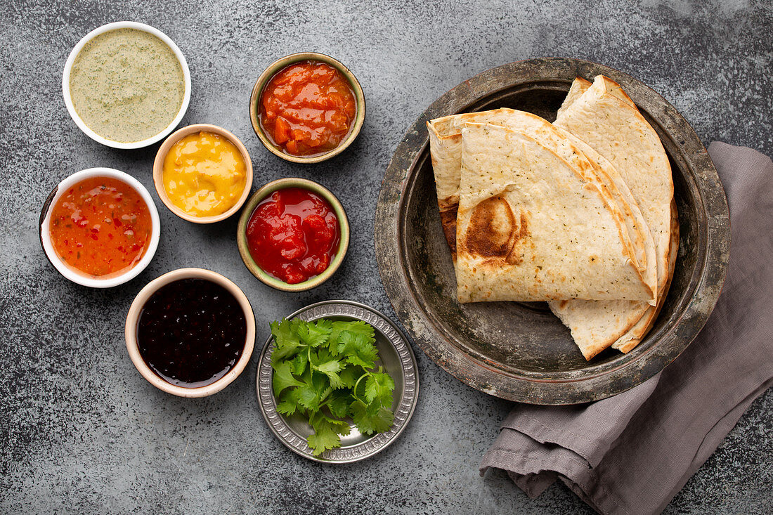 Assorted Indian chutneys in rustic bowls and flatbread on grey concrete background