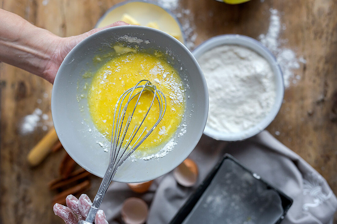 Whipping eggs in black bowl on wooden table with lemon, flour, butter and cinnamon sticks ingredients for cake
