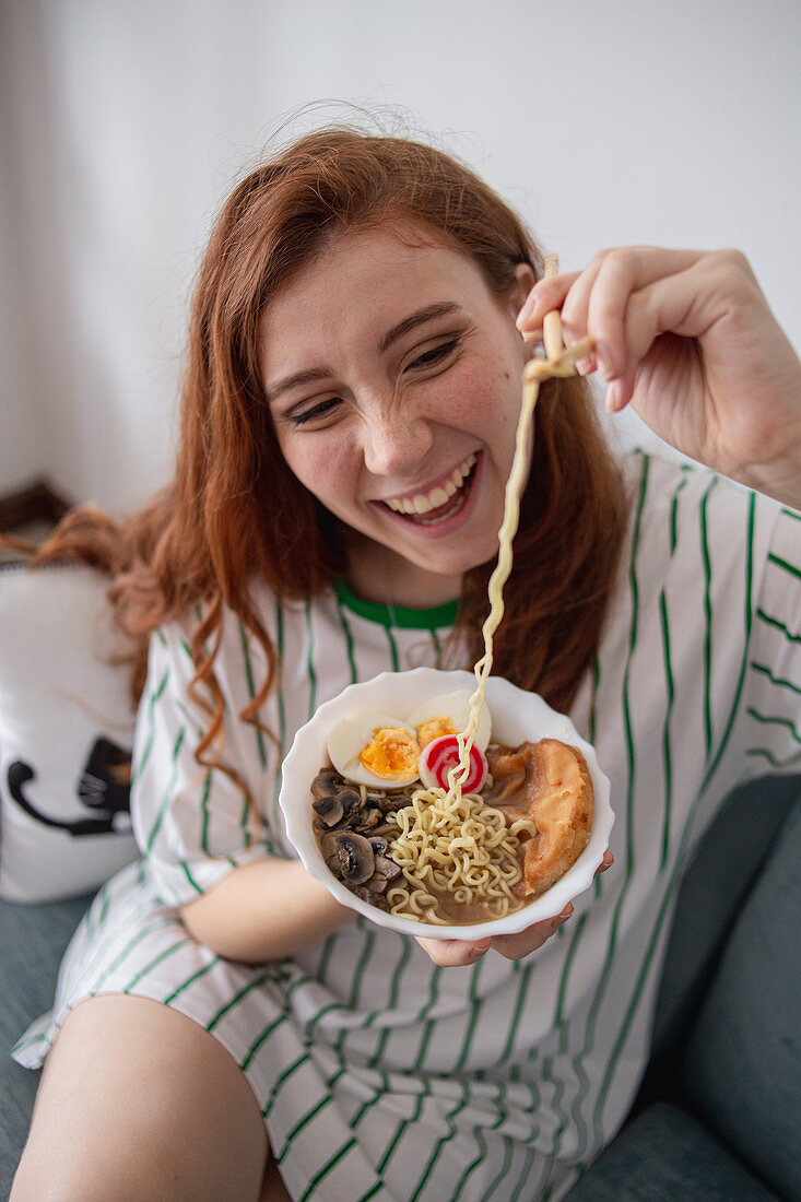 Cheerful redhead female laughing and picking noodles from bowl of tasty ramen while sitting on couch at home