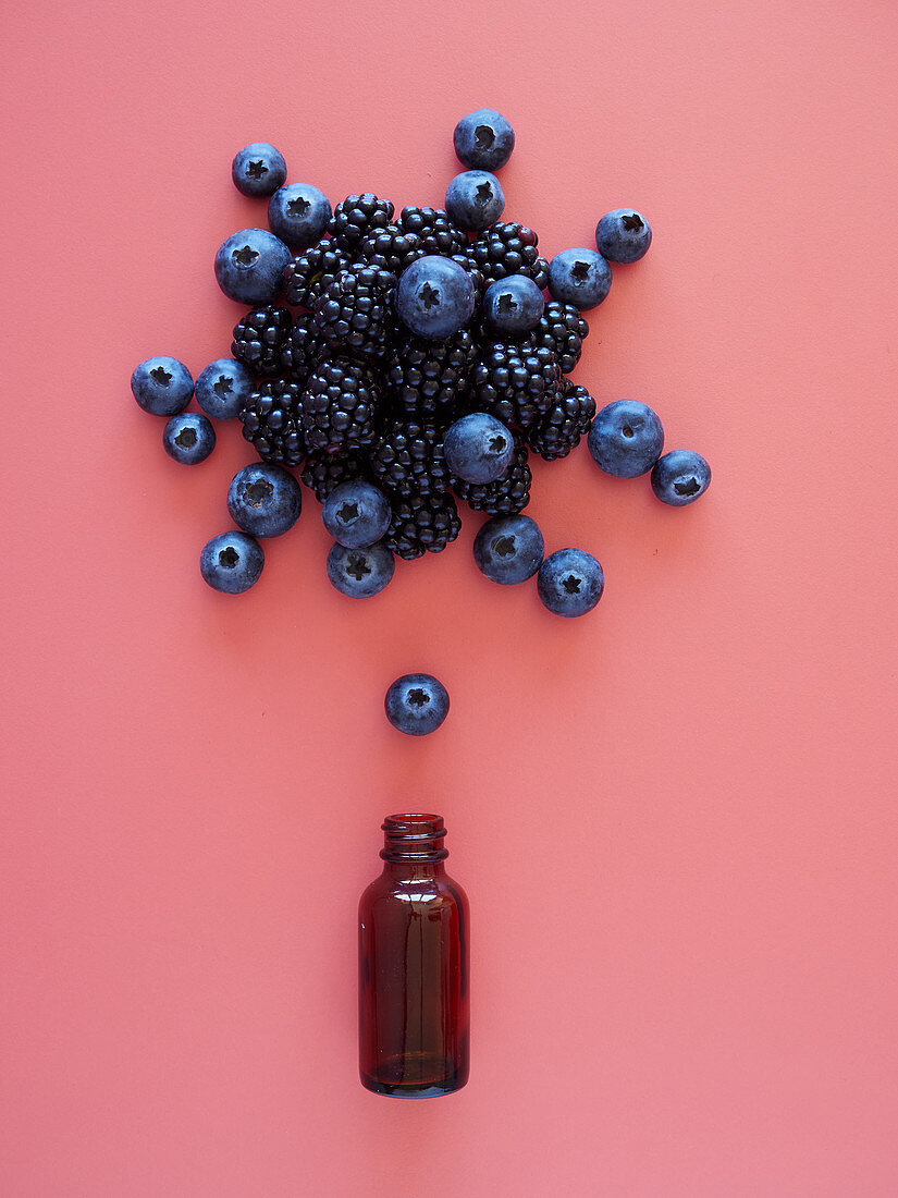 Top view composition of blueberries and blackberries arranged over small glass bottle on pink background