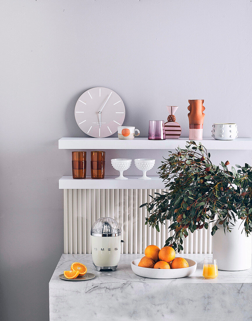 Marble counter with citrus press, oranges and leaf sprig in vase, shelves with clock and dishes in background