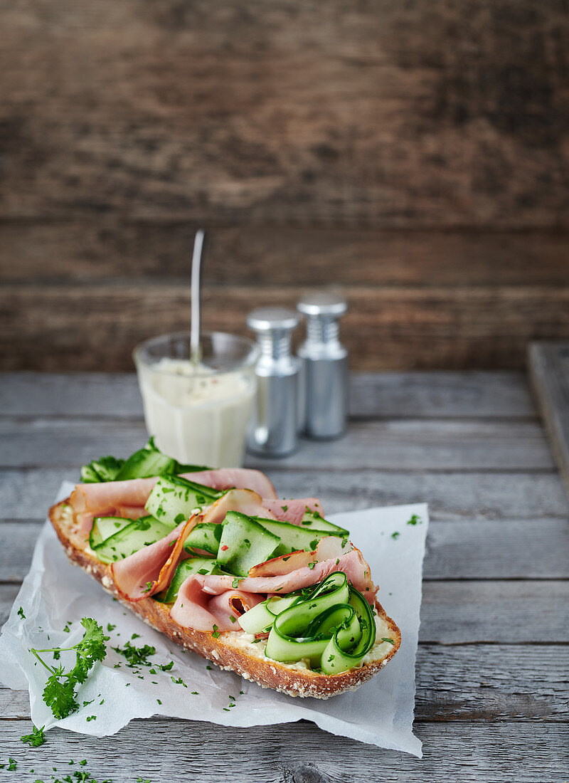 Baguette topped with cucumber and bacon on greaseproof paper