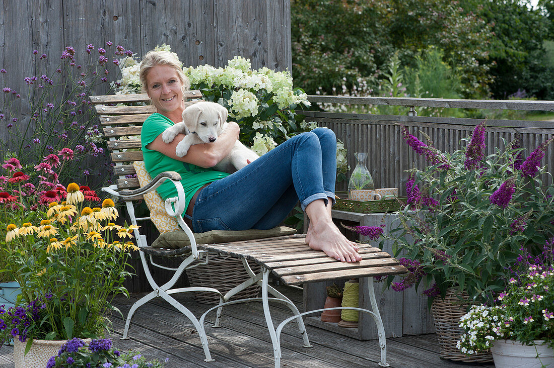 Balcony with Coneflower Conetto 'Banana' 'Raspberry' 'Butterfly Kisses', shrub hydrangea, verbena, summer lilac, woman on deck chair with dog Hera