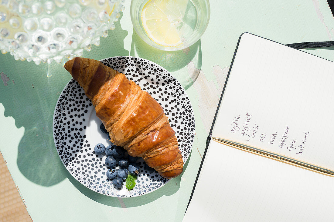 Teller mit Croissant und Blaubeeren neben aufgeschlagenem Notizbuch