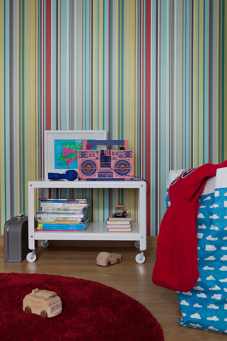 Side table with shelf on castors against multicoloured striped wallpaper in sibling's bedroom