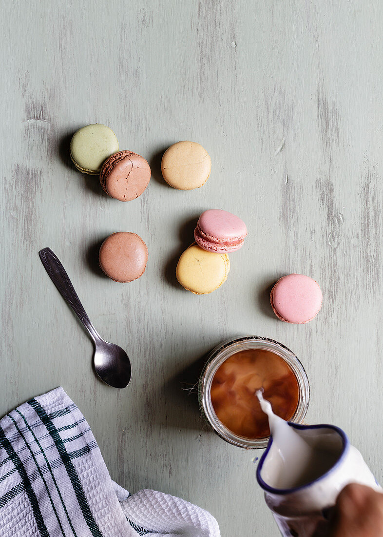 Person pouring milk into cup of coffee near bunch of colorful macaroons and spoon with napkin