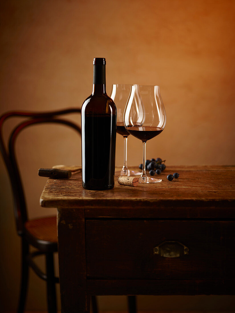 Rotweinstilleben mit Rotweinflasche, -glas und blauen Trauben auf Holztisch