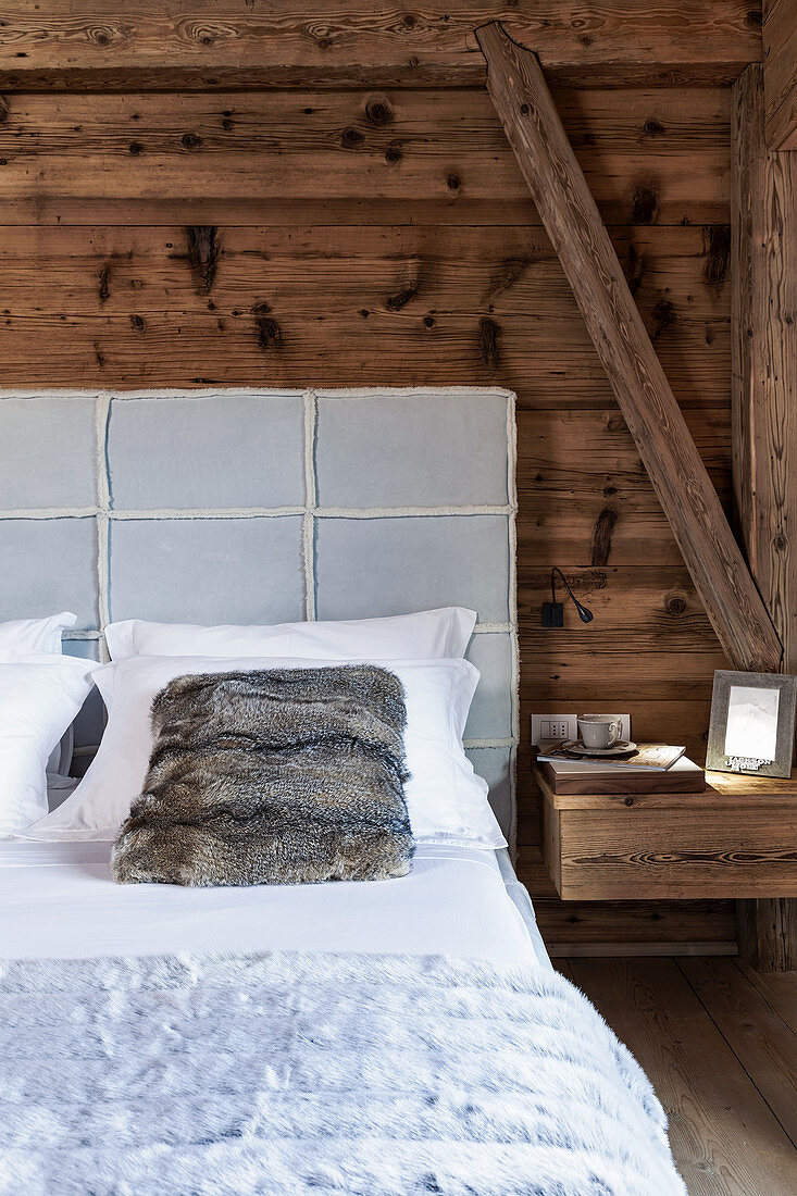 Bed with upholstered headboard and fur scatter cushion against rustic wooden wall