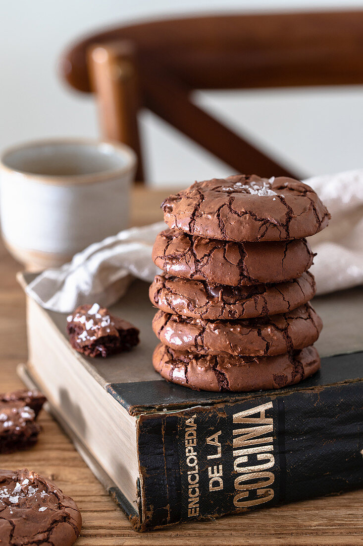 Stack of homemade chocolate brownie cookies and cookbook on wooden table against blurred background