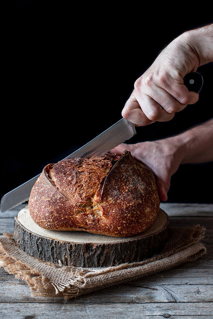 Unrecognizable person using knife to cut loaf of fresh sourdough bread on piece of wood against black background