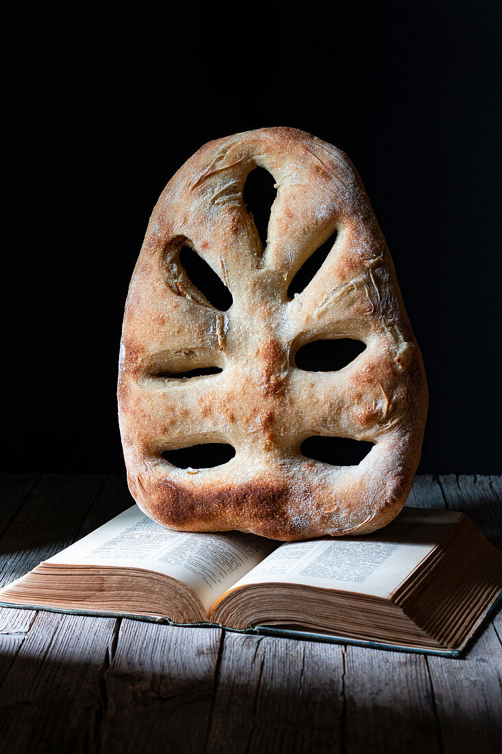 Loaf of fresh fougasse bread placed on open recipe book on wooden table against black background