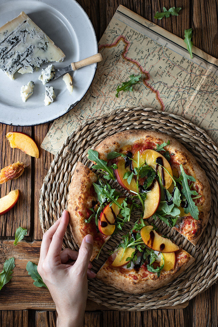 From above unrecognizable person taking piece of yummy summer pizza with peach and arugula near map and cheese on wooden table