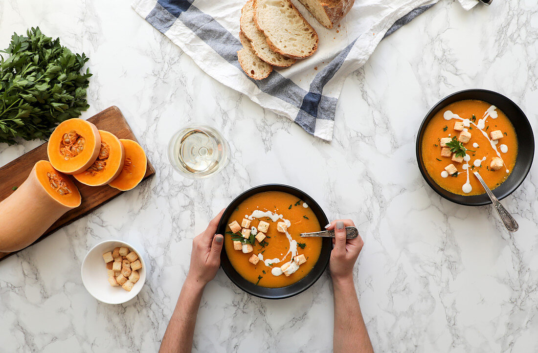 From above shot of unrecognizable person holding a bowl of orange pumpkin soup in a marble table with vegetables arrangements