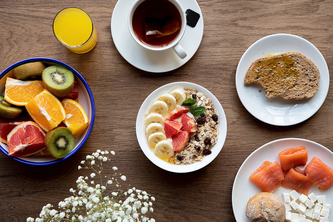 From above bowl of granola with tasty berries and banana served on table with plate of fruit and tea cup with for breakfast