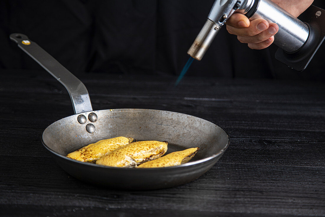 Crop person directing burner to pan with juicy endive preparing in frying pan on gray background