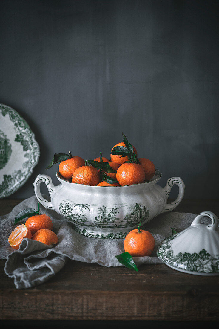 Sophisticated still life with ripe orange tangerines with green leaves in deep white bowl on table on gray background