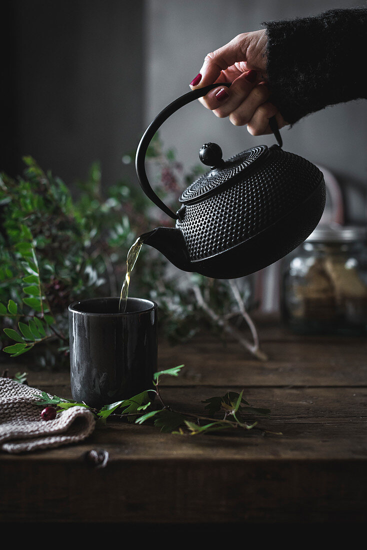 Round black teapot in hand of crop person pouring chamomile tea in black mug on wooden table decorating with green leaves