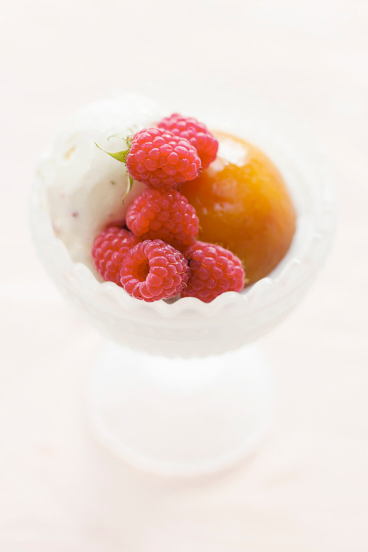 Frosted glass dessert bowl with a scoop of vanilla ice cream, a poached whole peach and freshly picked raspberries