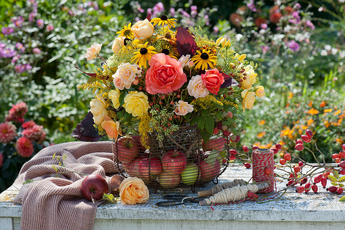 Late summer bouquet of roses, coneflowers, fennel, goldenrod, rose hips, and raspberries in a wire basket with apples
