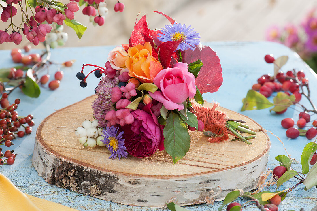 Kleiner Herbststrauß auf Holzscheibe: Rosen, Astern, wilder Wein, Fetthenne, Pfaffenhütchen und Knallerbsen