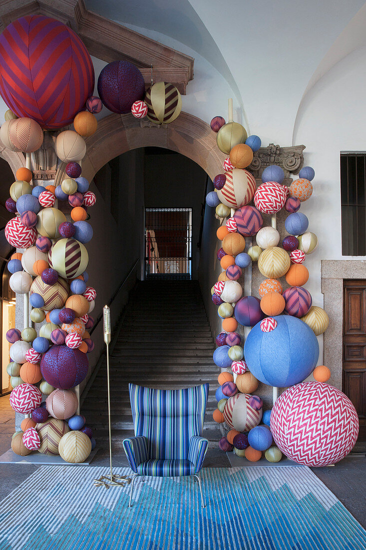 Blue armchair on rug at foot of historic stairs with arched doorway decorated with multicoloured fabric balls