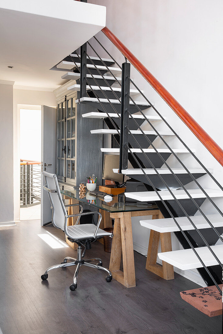 Glass desk on wooden trestles below modern open staircase