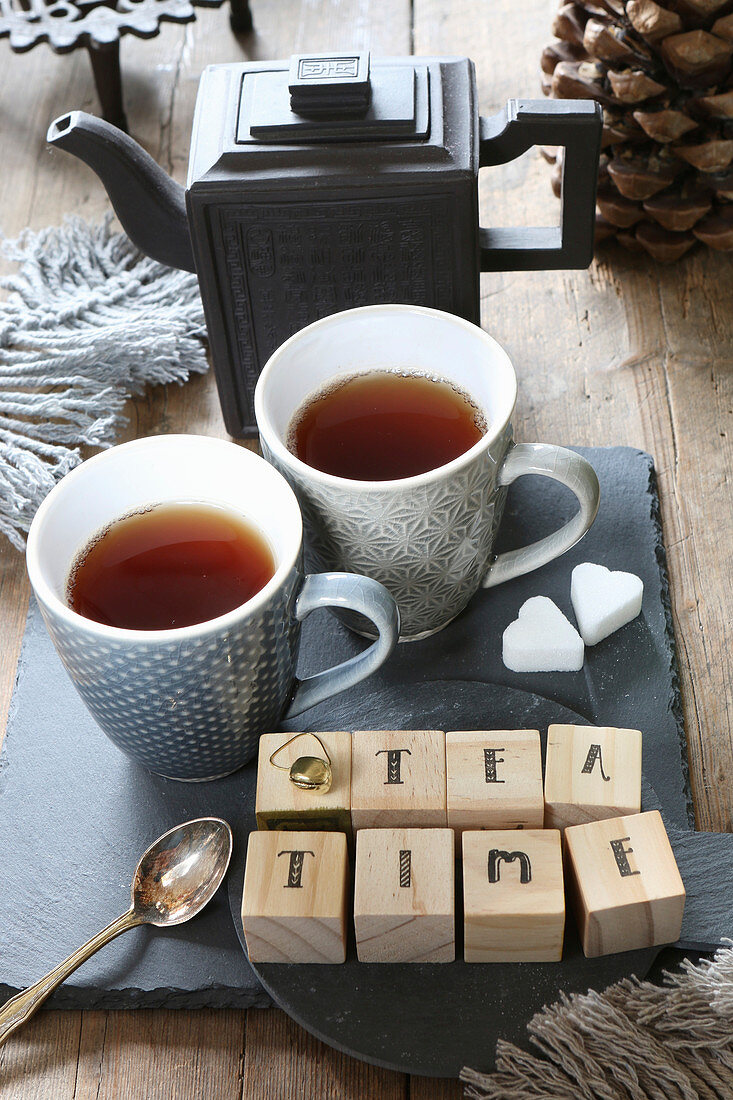 Wooden cubes with lettering spelling 'Tea time', teapot and teacups on slate