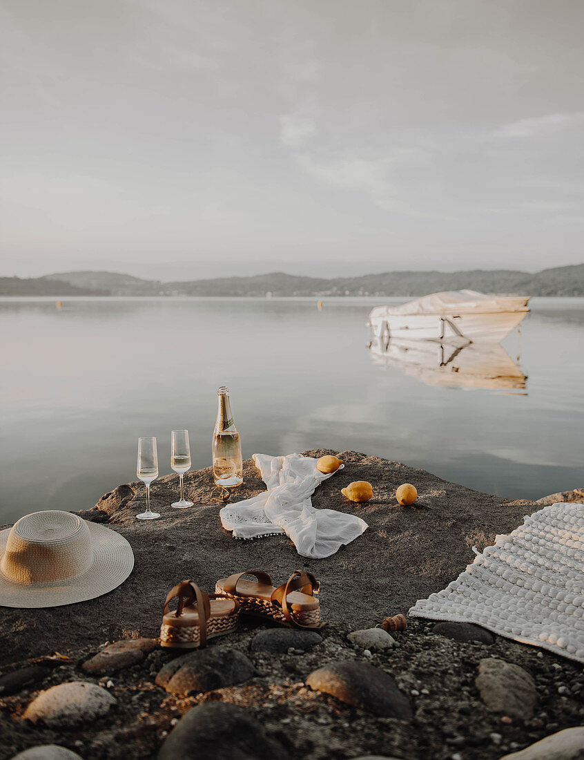 Bottle of sparkling wine, glasses, sunhat, sandals and blanket on rocky lake shore