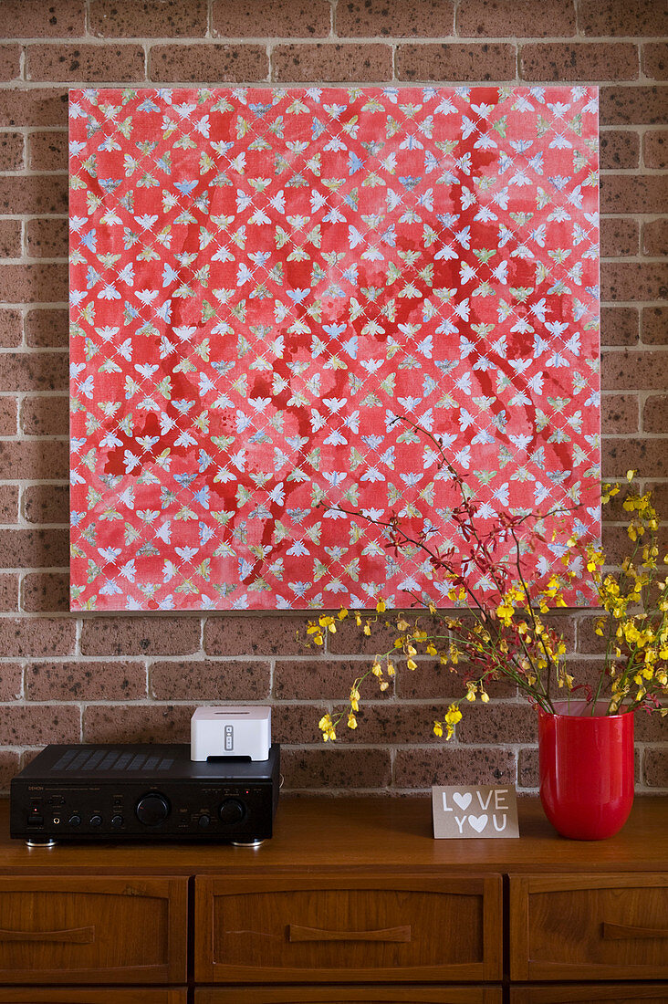 Wandbild mit rotem geometrischem Muster an Ziegelwand über Holzkommode