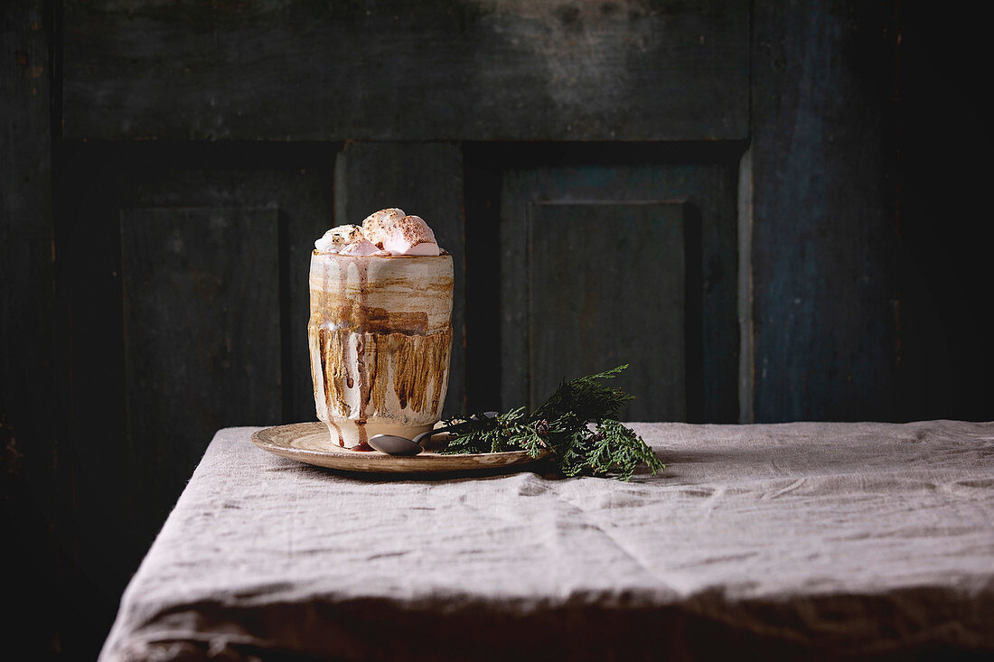 Hot chocolate or cocoa with marshmallow served in ceramic mug with saucer and thuja branch on grey table cloth