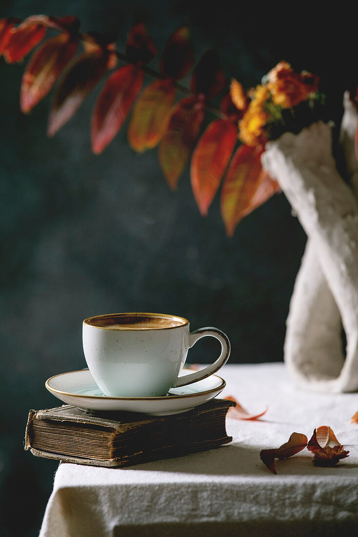 Cup of black coffee espresso standing on white table cloth in dark room with autumn leaves and flowers in clay vase, old books