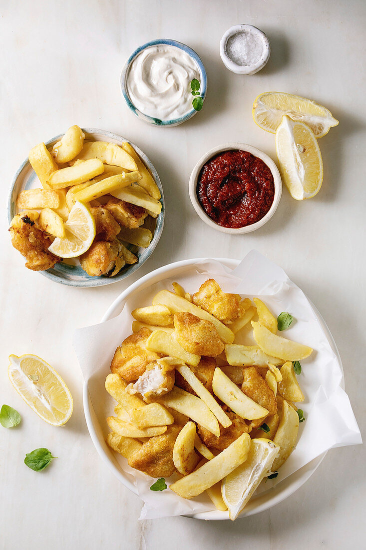 Classic british fast food fish and chips served in frying basket with lemons, red and white sauce, salt in ceramic bowls over wooden plank background