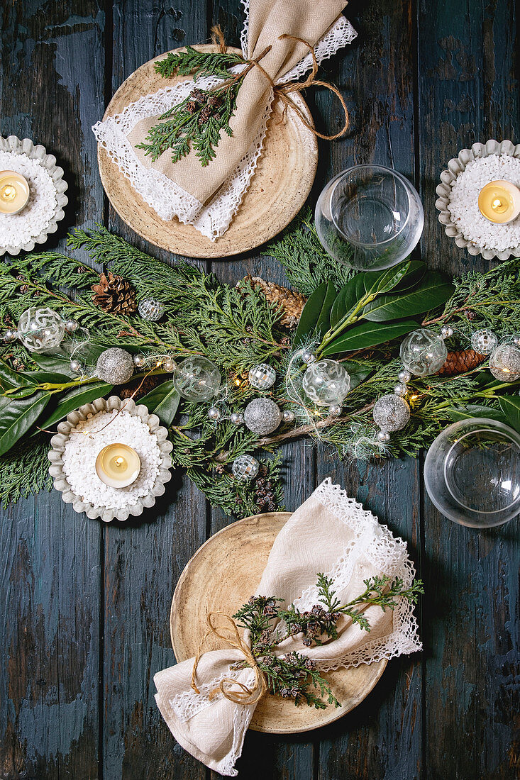 Christmas or New year table setting with empty ceramic plates, glasses, napkins, Christmas thuja wreath, luminous garland and burning candles
