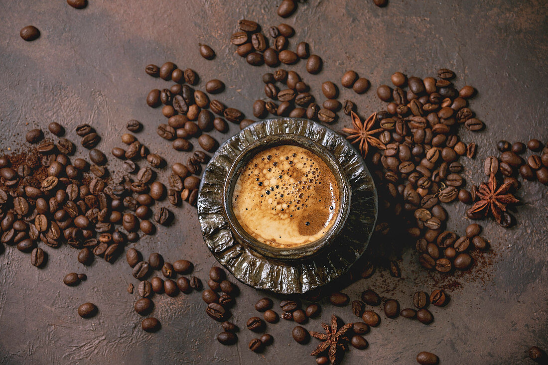 Black coffee espresso with foam in black ceramic cup, with saucer and roasted beans above over brown texture background