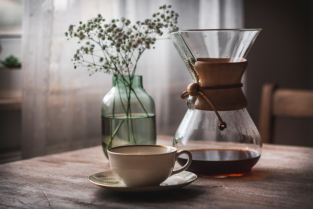 A chemex carafe, a coffee cup and a glass vase on a wooden table