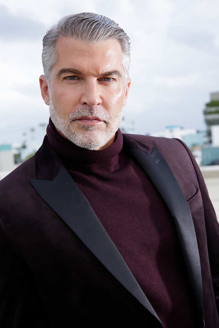 A grey-haired man on a terrace wearing a dark-red turtleneck jumper and a jacket