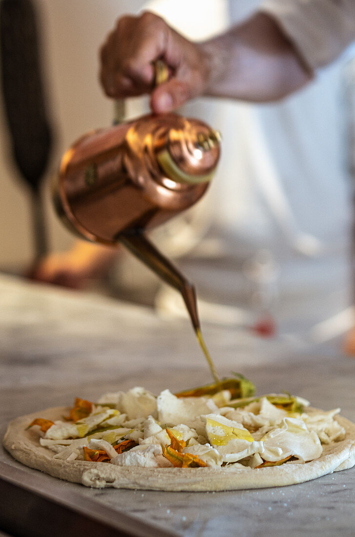 Crop anonymous Italian male chef pouring oil from metal teapot onto round pizza prepared for baking