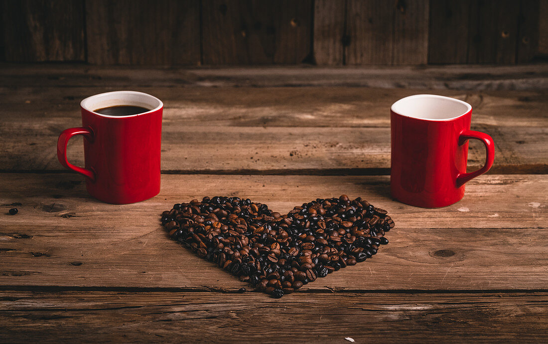 Ceramic mugs with hot drink placed on wooden table with grains arranged in shape of heart