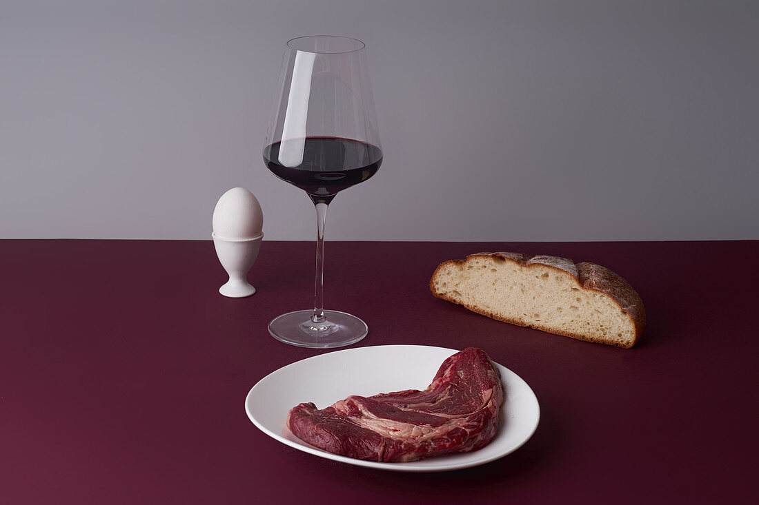 Stillleben mit Rotweinglas, Rindersteak, Brot und Ei