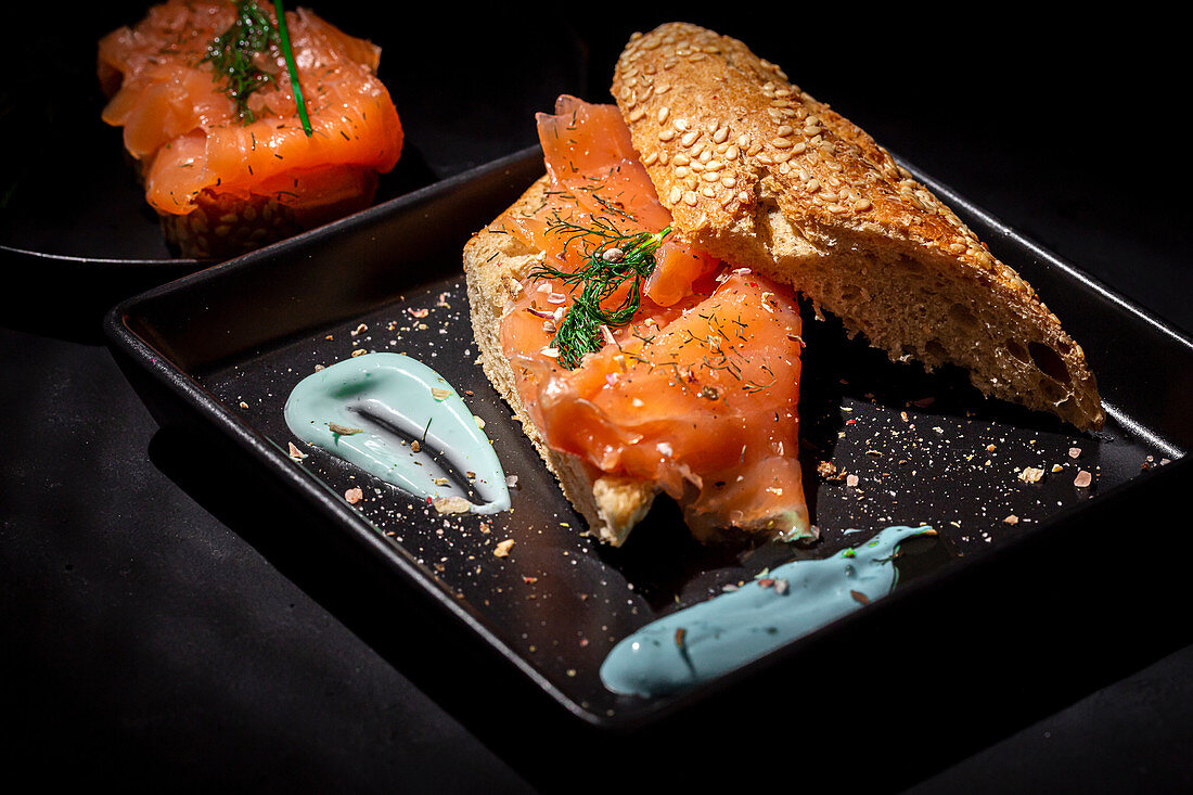 Smoked salmon and fennel herb on top of seeded bread on a dark plate and dark table