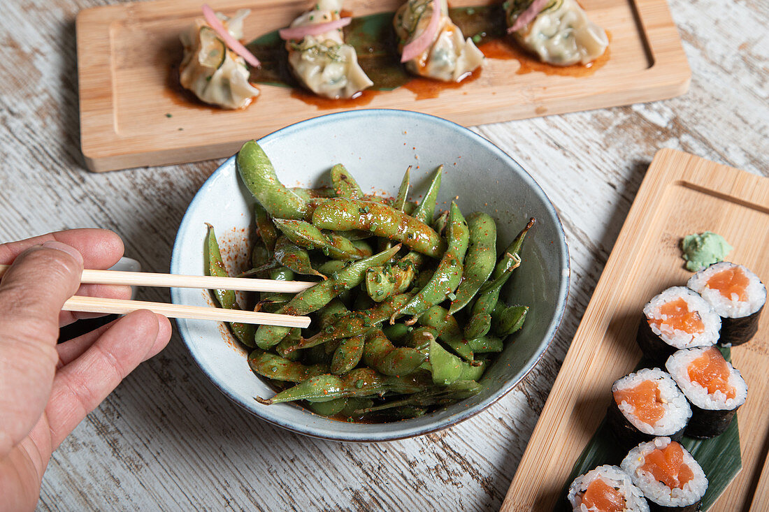 Bowl with edamame soy beans in pods served on wooden table with sushi rolls and boiled gyoza dumplings