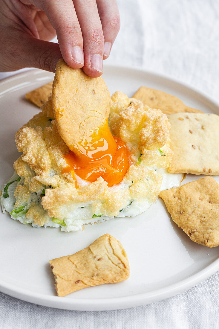 Hand dipping cracker in cloud eggs with green vegetables