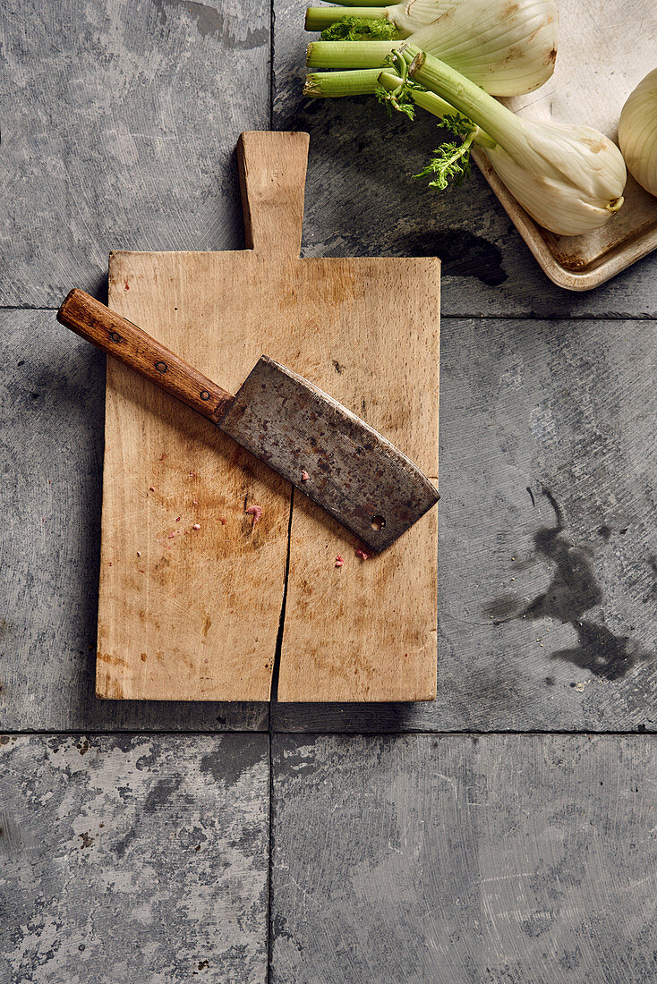 A meat cleaver on a chopping board next to fennel bulbs