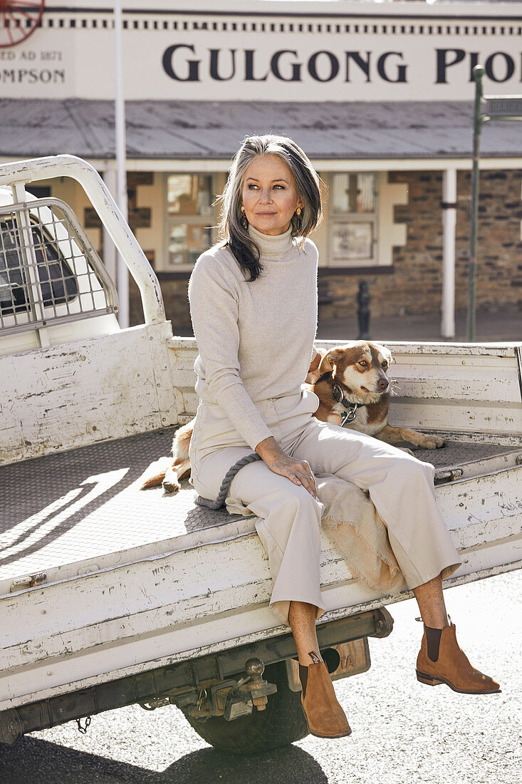 A grey-haired woman wearing a turtleneck jumper and light trouser sitting on a pick-up truck with a dog