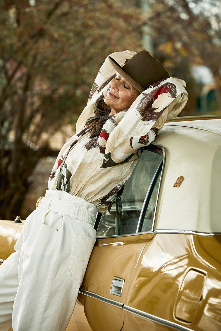A grey-haired woman wearing a hat, a blouse and white trousers leaning against a classic car