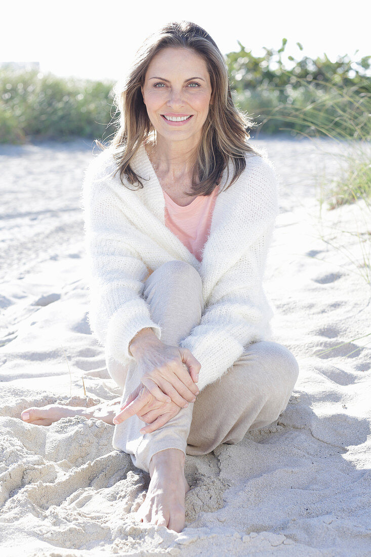 A long-haired woman sitting in the sand on the beach wearing a light jumper and trousers