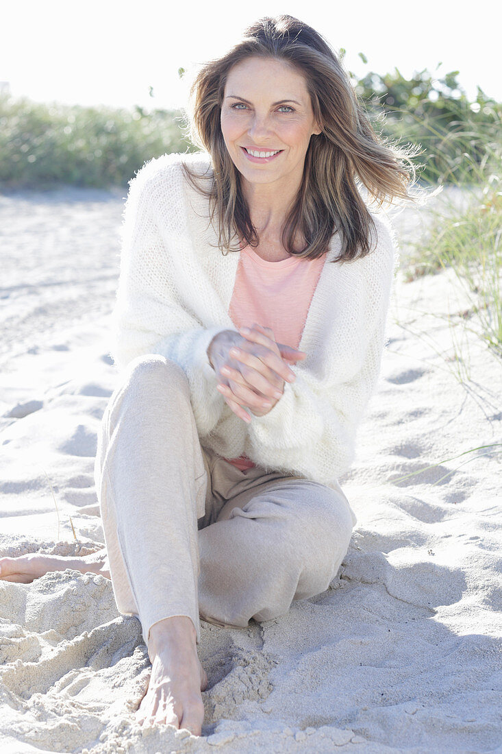 A long-haired woman sitting in the sand on the beach wearing a light jumper and trousers