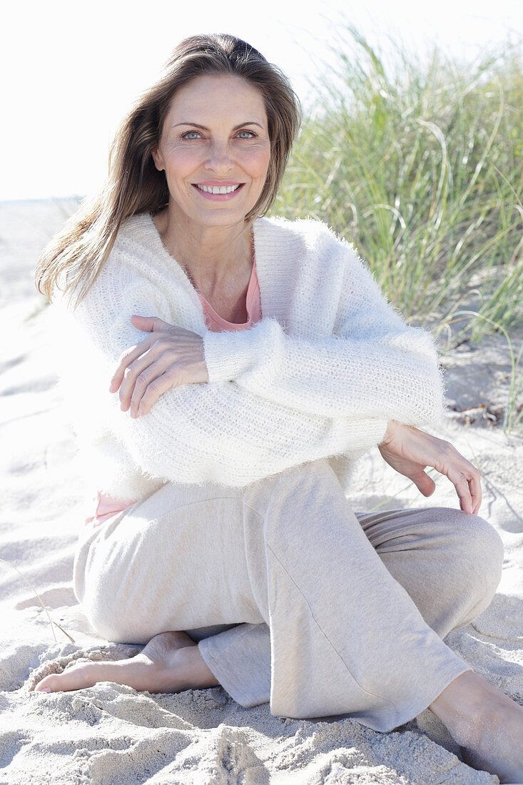 A long-haired woman sitting in the sand on the beach wearing a light jumper and trousers