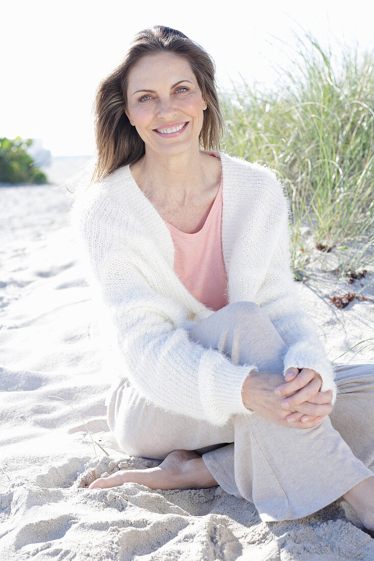 A long-haired woman sitting in the sand on the beach wearing a light jumper and trousers